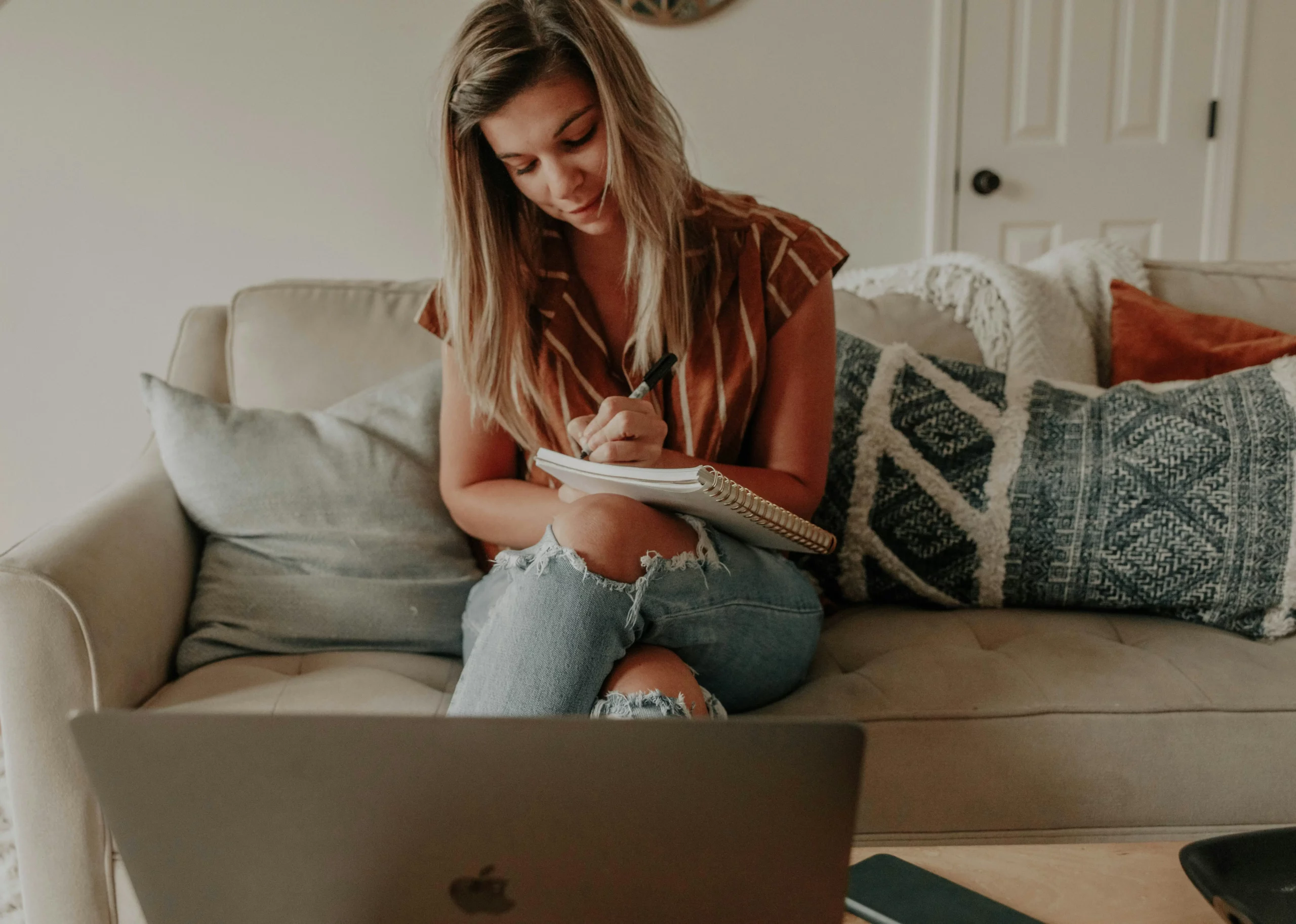 Une femme suit une formation en ligne depuis son canapé. Elle prend des notes et a son ordinateur devant elle.